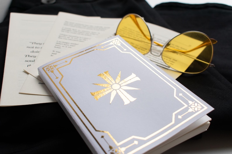 May include: A small, light gray book with gold accents and a gold sunburst design. Yellow-tinted sunglasses rest on top of the book and pages of text. The items are on a black surface.