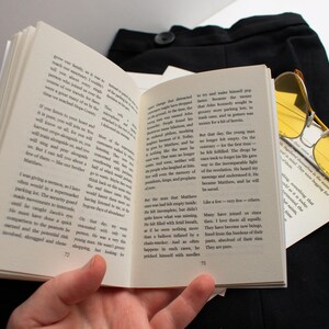 May include: An open book with text on the pages, held by a hand. Yellow-tinted sunglasses rest on the book, with black fabric in the background. The book pages are numbered 72 and 73.