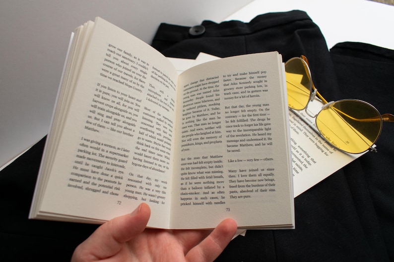 May include: An open book with text on the pages, held by a hand. Yellow-tinted sunglasses rest on the book, with black fabric in the background. The book pages are numbered 72 and 73.