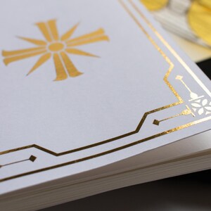 May include: Close-up of a stack of white paper with a gold foil design. The design includes a central sunburst and geometric border. The paper is stacked with the top sheet showing the design.