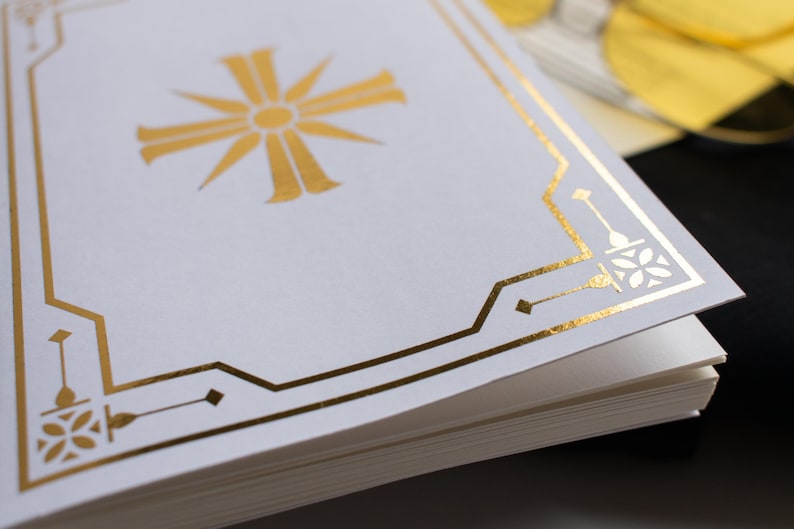 May include: Close-up of a stack of white paper with a gold foil design. The design includes a central sunburst and geometric border. The paper is stacked with the top sheet showing the design.