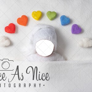 K&ouml;nnte beinhalten: Ein neugeborenes Baby, das eine wei&szlig;e Strickm&uuml;tze und einen wei&szlig;en Strickpullover tr&auml;gt, liegt auf einer wei&szlig;en Decke. Ein Regenbogen aus Filzherzen ist in einem Bogen &uuml;ber dem Kopf des Babys angeordnet. Das Gesicht des Babys ist nicht sichtbar. Der Text "Twice As Nice Photography" ist in der linken unteren Ecke des Bildes sichtbar.
