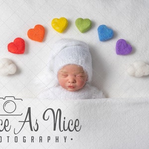 K&ouml;nnte beinhalten: Ein neugeborenes Baby, das eine wei&szlig;e Strickm&uuml;tze und ein wei&szlig;es Strickoutfit tr&auml;gt, schl&auml;ft auf einer wei&szlig;en Decke. Ein Regenbogen aus Filzherzen ist in einem Bogen &uuml;ber dem Kopf des Babys angeordnet. Die Worte "Twice As Nice Photography" sind unter dem Baby gedruckt.