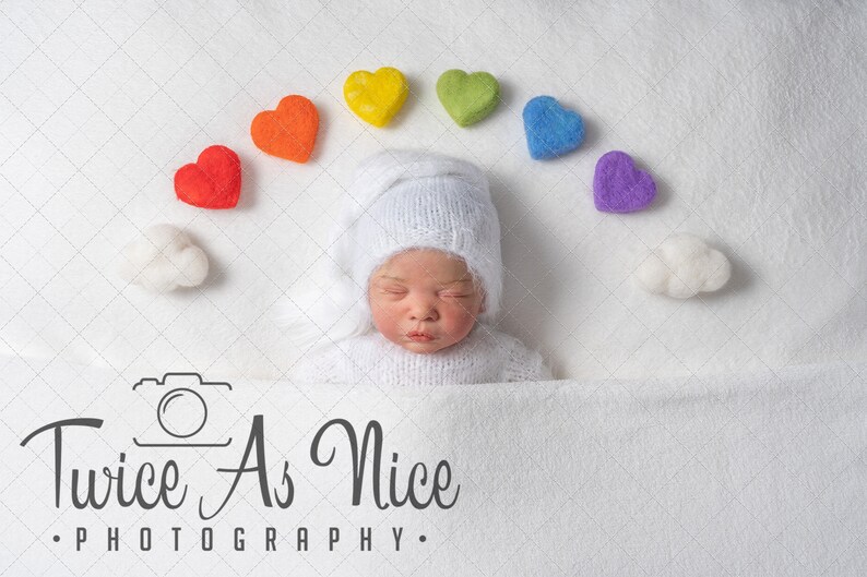 K&ouml;nnte beinhalten: Ein neugeborenes Baby, das eine wei&szlig;e Strickm&uuml;tze und ein wei&szlig;es Strickoutfit tr&auml;gt, schl&auml;ft auf einer wei&szlig;en Decke. Ein Regenbogen aus Filzherzen ist in einem Bogen &uuml;ber dem Kopf des Babys angeordnet. Die Worte "Twice As Nice Photography" sind unter dem Baby gedruckt.