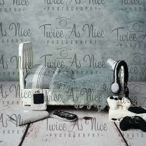 Puede incluir: Una cama blanca en miniatura con una manta gris y blanca, una pequeña televisión, un mando a distancia, unos auriculares y un mando de videojuegos. La cama está sobre una superficie de madera blanca con el texto "Twice As Nice Photography" repetido en todo el fondo.