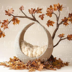 May include: A white wicker basket shaped like a crescent moon, with a white fluffy blanket inside, sits on a bed of brown autumn leaves. The basket is suspended from a branch with brown leaves.