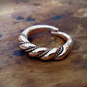 May include: A bronze-colored ring with a braided design. The ring has an open design, allowing for size adjustment. The braided section is centered on the ring, with smooth, rounded bands on either side. The ring is set against a wooden surface.