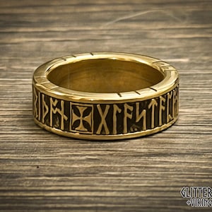May include: A gold-colored ring with engraved runic symbols and a cross design. The ring is set against a wooden background. The ring is a Viking-themed piece.
