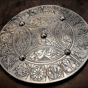 May include: A circular silver metal shield with intricate Celtic knotwork designs and figures. The shield has a raised center and is decorated with a series of concentric circles.