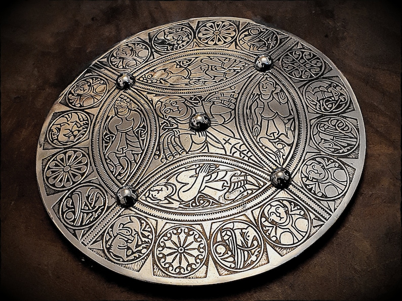 May include: A circular silver metal shield with intricate Celtic knotwork designs and figures. The shield has a raised center and is decorated with a series of concentric circles.