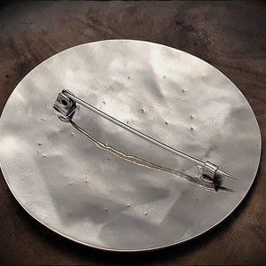 May include: The back of a silver circular brooch with a pin and catch. The brooch has a textured surface.