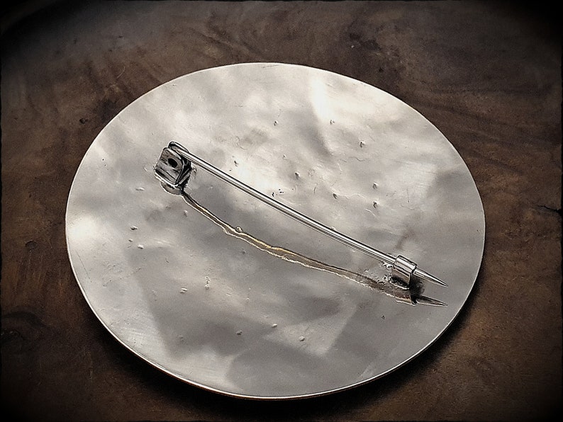 May include: The back of a silver circular brooch with a pin and catch. The brooch has a textured surface.