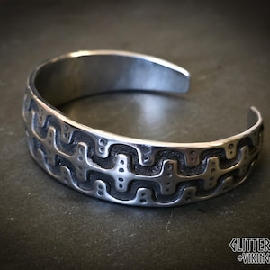 May include: A silver-toned metal cuff bracelet featuring a repeating embossed geometric pattern with a dark, recessed background. The bracelet has an open-ended design and includes the text GLITTERBEARD'S VIKING CRAFTS in the bottom right corner.