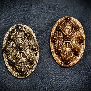 Può includere: Due spille ovali in metallo dorato con disegni intricati che presentano teschi e ossa incrociate. Le spille hanno una finitura testurizzata e invecchiata.