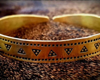 May include: A gold-toned metal bracelet with a decorative pattern of triangles and dots. The bracelet is on a brown fur background.