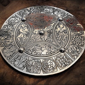 May include: A silver circular disc with intricate Celtic knotwork designs and four raised metal studs. The design features a central motif of four figures within a circular frame.