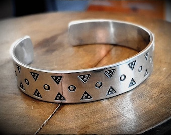 May include: A silver cuff bracelet with a geometric pattern of triangles and circles. The bracelet is made of a shiny metal and has an open design. The triangles contain three small dots. The bracelet is approximately 2.5 inches wide.