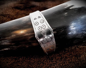 May include: A silver metal band with a decorative pattern of circles and triangles. The band is attached to a dark brown object.