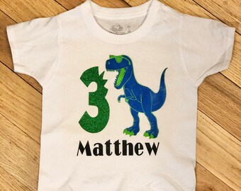 May include: White t-shirt with a green number 3 and a blue dinosaur wearing sunglasses. The text "Matthew" is printed below the dinosaur.