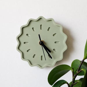 May include: A sage green wall clock with a scalloped, wavy edge and a circular face. The clock features twelve recessed dash-shaped hour markers, a central brass-colored hub, and three thin black hands indicating the time on the dial.