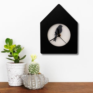 Puede incluir: Un reloj de pared negro con forma de casa, con la silueta de un pájaro sobre un fondo blanco. El reloj tiene manecillas negras y está montado en una pared blanca. Plantas en macetas sobre una superficie de madera.