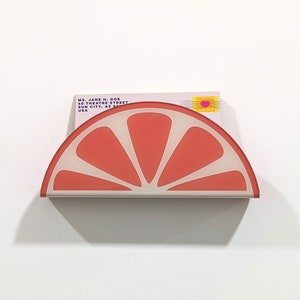 May include: A pink and white ceramic half-grapefruit mail holder with a white background. The mail holder is shaped like a half-grapefruit with a white background and a pink grapefruit slice on top. The mail holder is designed to hold mail and other small items.