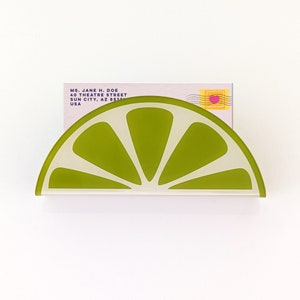 May include: A green lime-shaped ceramic holder for business cards. The holder is shaped like a slice of lime with a white background and green segments. The holder is sitting on a white surface.