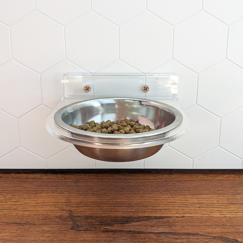 May include: A clear acrylic pet food bowl holder mounted on a white tiled wall. The holder has a stainless steel bowl with dry pet food inside.