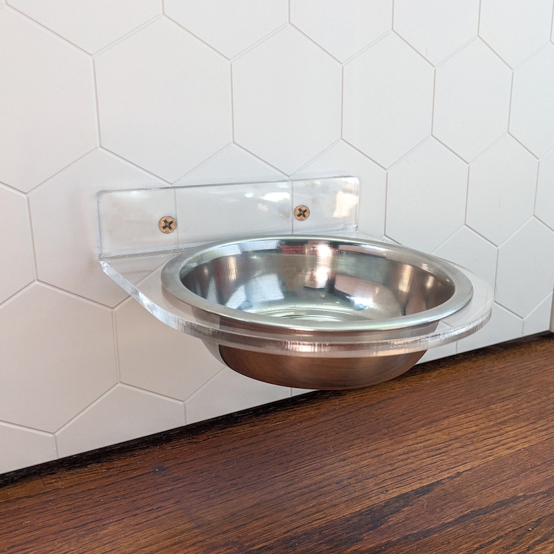 May include: A clear acrylic pet bowl holder mounted on a white hexagon tile wall. The holder has two screws and a stainless steel bowl.