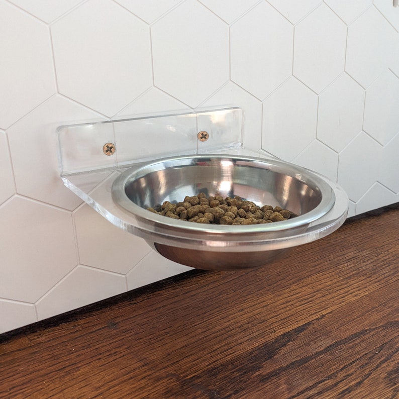 May include: A clear acrylic pet bowl holder mounted on a white tiled wall. A stainless steel bowl filled with dry pet food is attached to the holder.