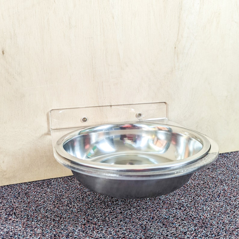 May include: A stainless steel pet bowl mounted to a wall with a clear plastic bracket. The bowl is empty and ready for food or water.