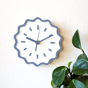 May include: A round wall clock with a white face and a wavy, light blue border. The clock has black hands and blue dashes marking the hours. The clock is mounted on a white wall, with green plant leaves in the corner.