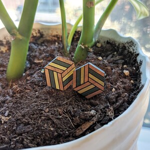 May include: A pair of wooden cufflinks with a geometric design in shades of green, yellow, and brown. The cufflinks are in focus, and the background is blurred.