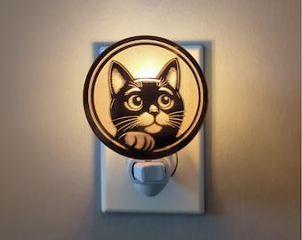 May include: A black cat night light with a circular design. The cat's face is centered, with large eyes and a paw visible. The night light plugs into a wall outlet and emits a soft glow.