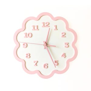 May include: A pink cloud-shaped wall clock with a white face. The clock has pink numerals and hands, and a scalloped edge. The clock's design is simple and modern, suitable for a nursery or child's room.