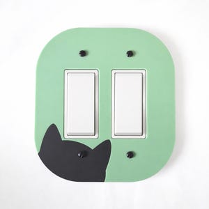 Puede incluir: Cubierta de interruptor doble de color verde claro con un diseño de gato negro. La cubierta tiene dos interruptores de luz blancos y se asegura con cuatro tornillos negros. Un accesorio decorativo para el hogar.