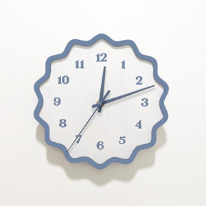 May include: A white clock with blue numbers and hands, set to 3:00. The clock has a wavy blue frame and is hanging on a white wall.