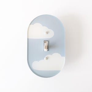 May include: An oval light switch plate featuring a light blue background with white cloud patterns. The plate includes a central toggle switch and two white mounting screws positioned above and below the switch mechanism.