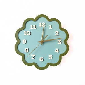 May include: A wall clock with a light blue face and a green scalloped edge. The clock has white numbers and gold-coloured hands. The clock is a decorative item for a home or office.