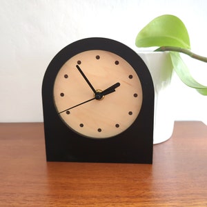 May include: A black wooden clock with a light wood face and black hands. The clock has a simple design with a rounded top and a square base.
