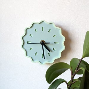 Puede incluir: Un reloj de pared verde claro con agujas negras y un borde ondulado y festoneado. El reloj está colgado en una pared blanca.