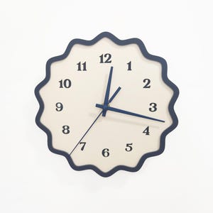 May include: A round wall clock with a cream-coloured face and navy blue hands and numbers. The clock has a navy blue, scalloped edge. The hands point to 3:15. The clock is a decorative item.