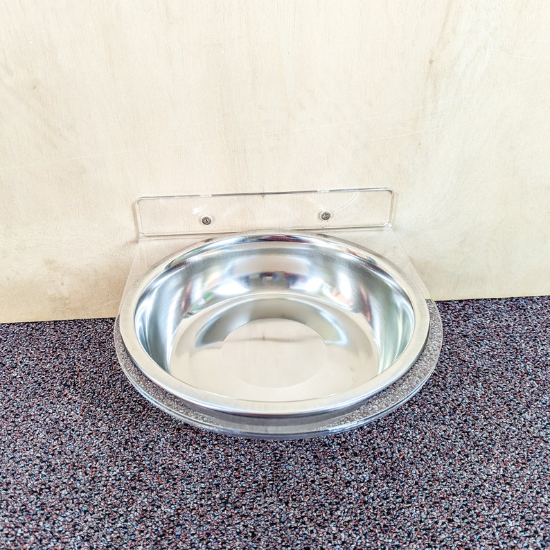 May include: A stainless steel pet bowl mounted to a wall with a clear plastic bracket. The bowl is empty and sits on a brown carpet.