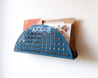 May include: A teal-colored, semi-circular wall-mounted mail organizer. The organizer has a woven pattern with circular cutouts. It holds several pieces of mail, including a brown paper envelope and postcards. The design is simple and modern.