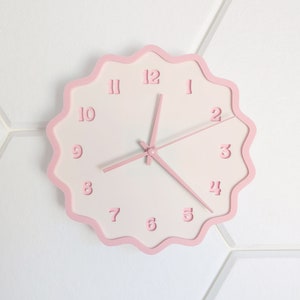 May include: A round wall clock with a scalloped pink frame and a white face. The clock features pink numerals and hands. The design is minimalist and modern, suitable for home decor.