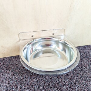 May include: A stainless steel pet bowl mounted to a wall with a clear plastic bracket. The bowl is empty and has a shiny, reflective surface.