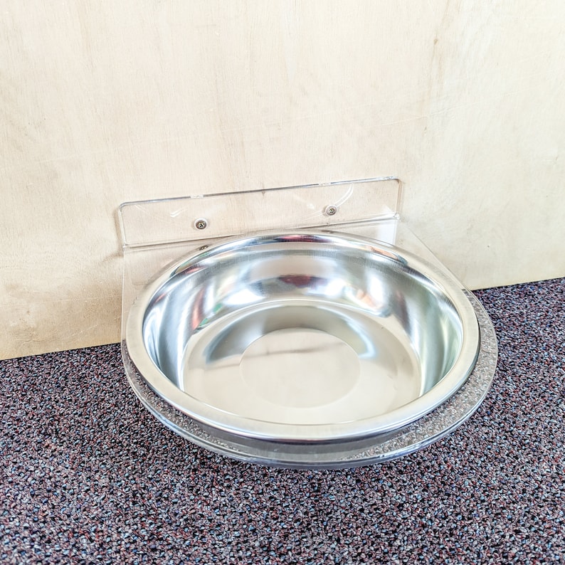 May include: A stainless steel pet bowl mounted to a wall with a clear plastic bracket. The bowl is empty and has a shiny, reflective surface.