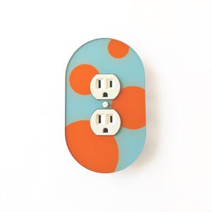 Puede incluir: Una placa de pared para enchufe eléctrico de forma ovalada con un fondo azul claro decorado con varios círculos naranjas sólidos de diferentes tamaños. La placa está centrada alrededor de un enchufe dúplex blanco estándar con un tornillo.