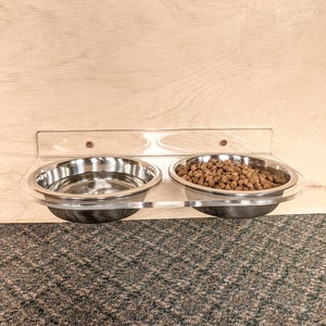 Pu&ograve; includere: Un porta ciotole per cibo e acqua per animali domestici in acrilico trasparente da parete con due ciotole in acciaio inox. Una ciotola &egrave; piena di cibo per animali domestici marrone.