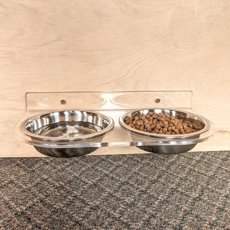 Pu&ograve; includere: Un porta ciotole per cibo e acqua per animali domestici in acrilico trasparente da parete con due ciotole in acciaio inox. Una ciotola &egrave; piena di cibo per animali domestici marrone.
